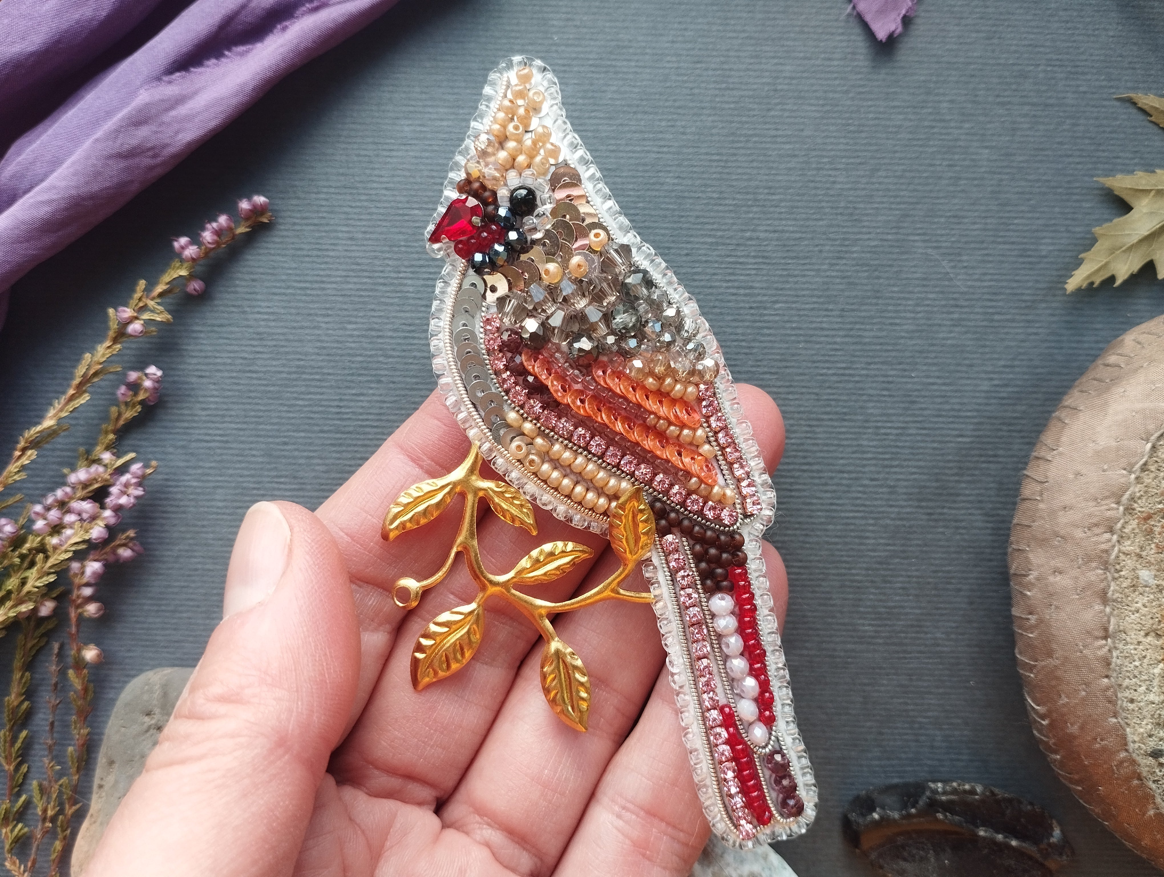 A hand holding a decorative bird brooch made of colorful beads and leaves.